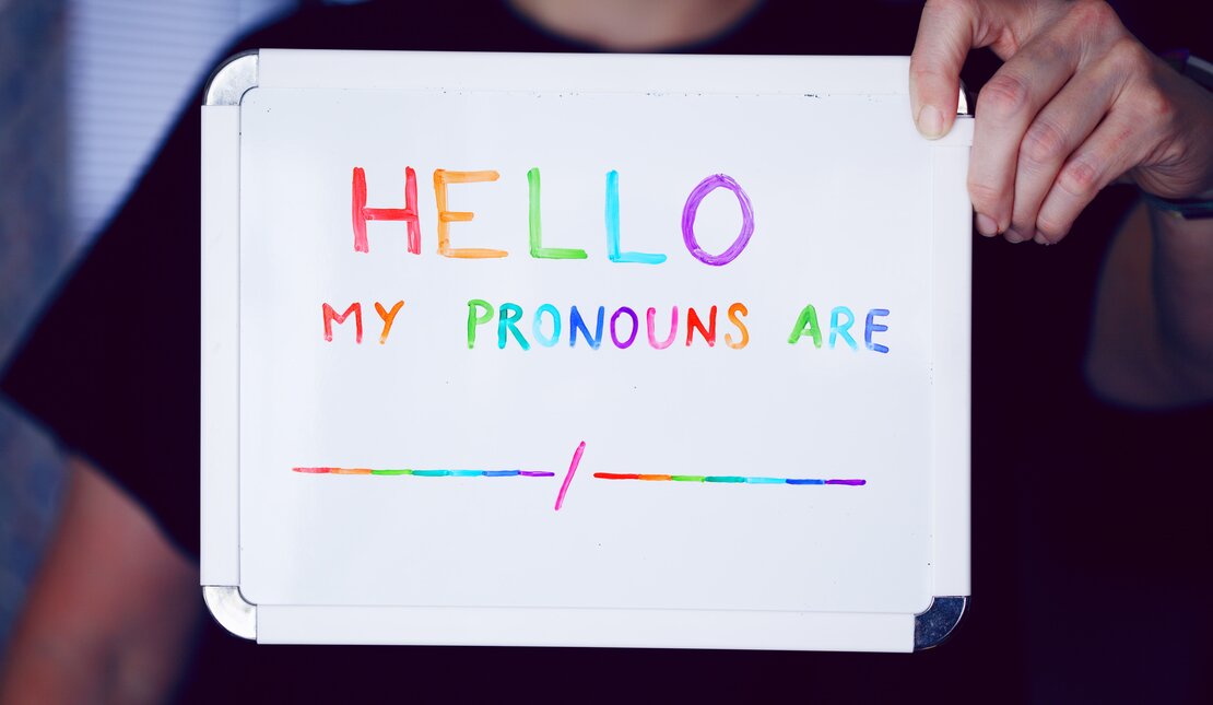 Person holding up a whiteboard wearing a black t-shirt. In rainbow marker, the sign reads hello, my pronouns are blank space slash blank space.