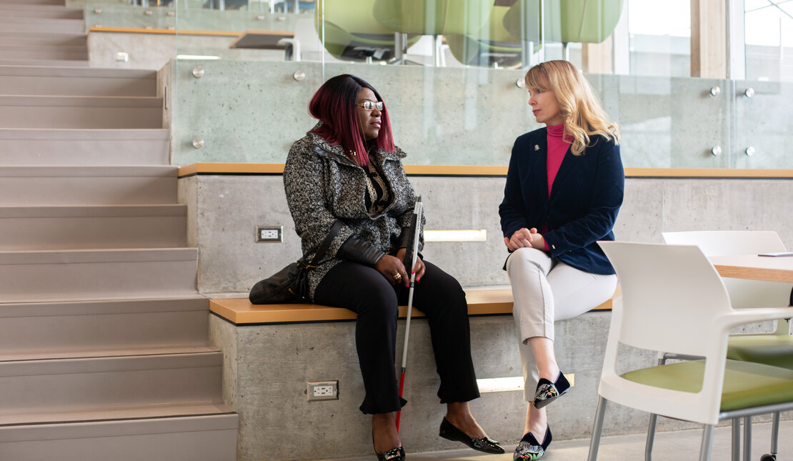 Laetitia Mfamobani, who has long pink and black hair and is wearing a white and black coat sitting beside Rebecca Blissett, who has long blonde hair and is wearing a pink shirt and navy blazer. The two are sitting on stair seats, Laetitia has a white cane.