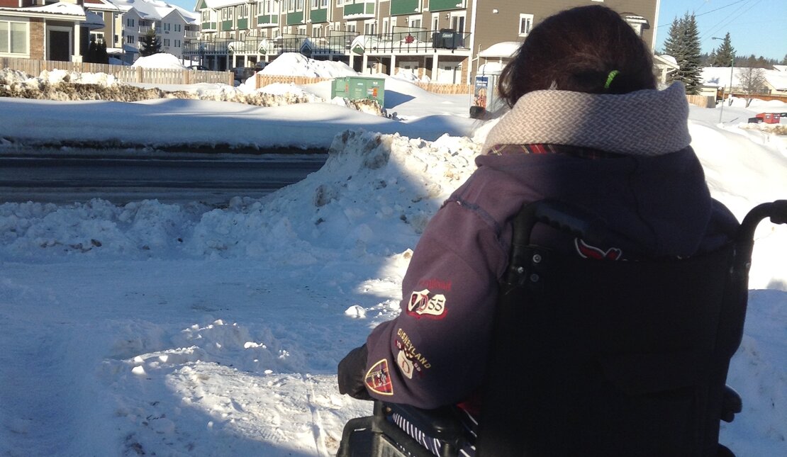 Back of person using a wheelchair wearing a purple winter jacket. It is winter with large piles of snow on the ground and apartment buildings in the distance. 