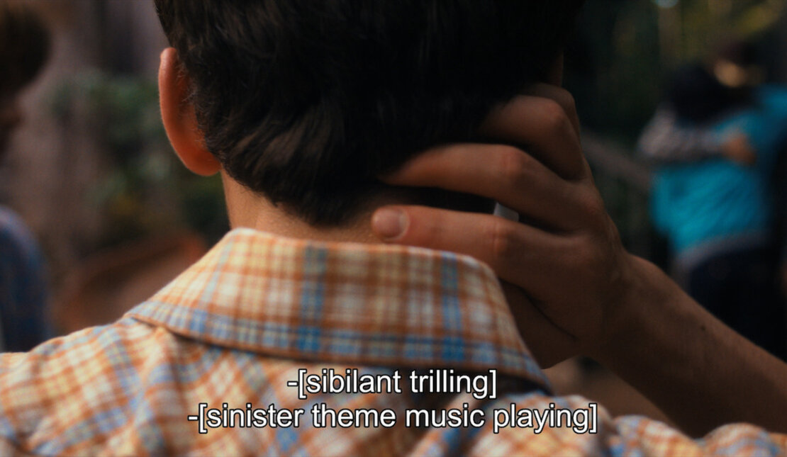 Back of the head of someone with short dark hair wearing a plaid shirt whose hand is on the back of their neck. Closed captioning located at the bottom of the image reads "sibilant trilling" and "sinister theme music playing".