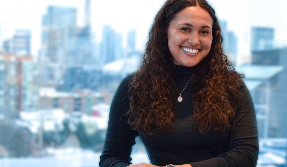 Haley Rae Dinnall-Atkinson, who is a woman with long brown curly hair smiling and leaning on a brown chair. She is wearing a black turtle neck sweater and blue jeans. Behind her is an open window to a city landscape.