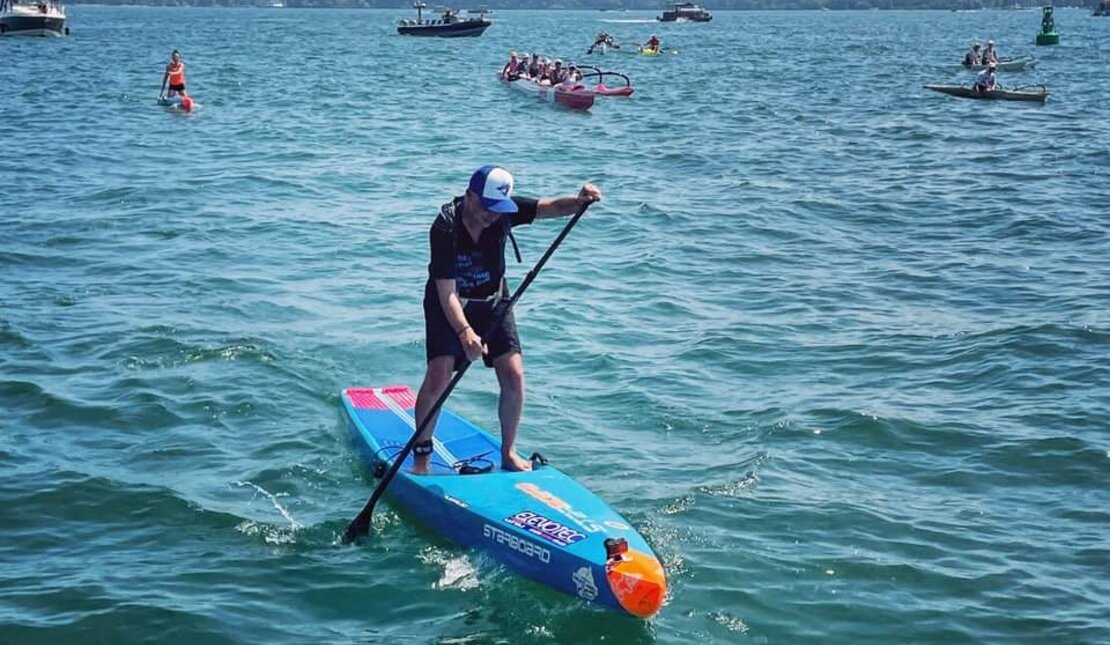 Mike Shoreman paddleboarding. He is wearing a black t-shirt and shorts and a Toronto Blue Jays baseball cap. His paddleboard is blue and orange. There are boats behind him.