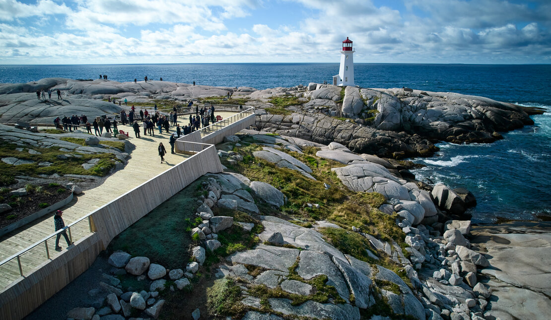 Aerial view of Peggy's Cove lighthouse. There is a large, wooden accessible viewing deck with a crowd of people towards the front. The viewing deck is built on top of large rocks. The lighthouse is red and white and overlooks the ocean.
