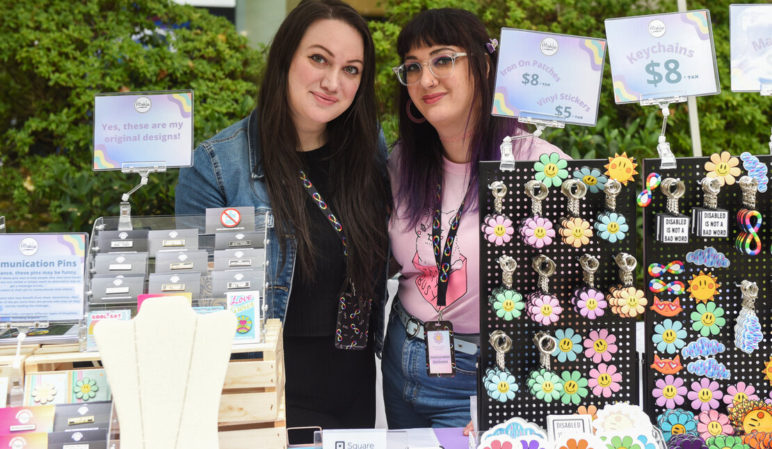 We Belong! Festival founder Margaux Wosk on the right with sister Becky Wosk. Margaux has long dark hair with purple in it, is wearing a pink t-shirt and has glasses. Becky has long dark hair and is wearing a black outfit with a denim jacket. They are in front of a stand of items that are for sale.