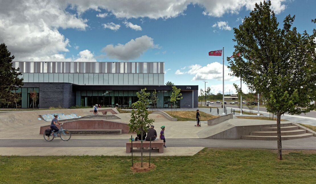 An outside view of Kingston East Community Centre. There are people outside using the accessible pathways and ramps, and benches. 