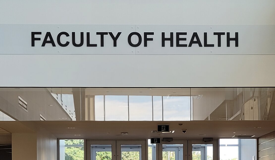 Inside the University of Waterloo. The Faculty of Health sign is above a set of doors with stairs and an accessible ramp.