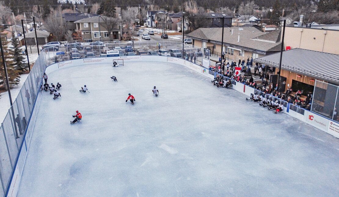 Aerial view of Parkdale ice rink. The rink is outdoors, and people are playing sledge hockey. There are houses and cars in the neighbourhood beside the arena.