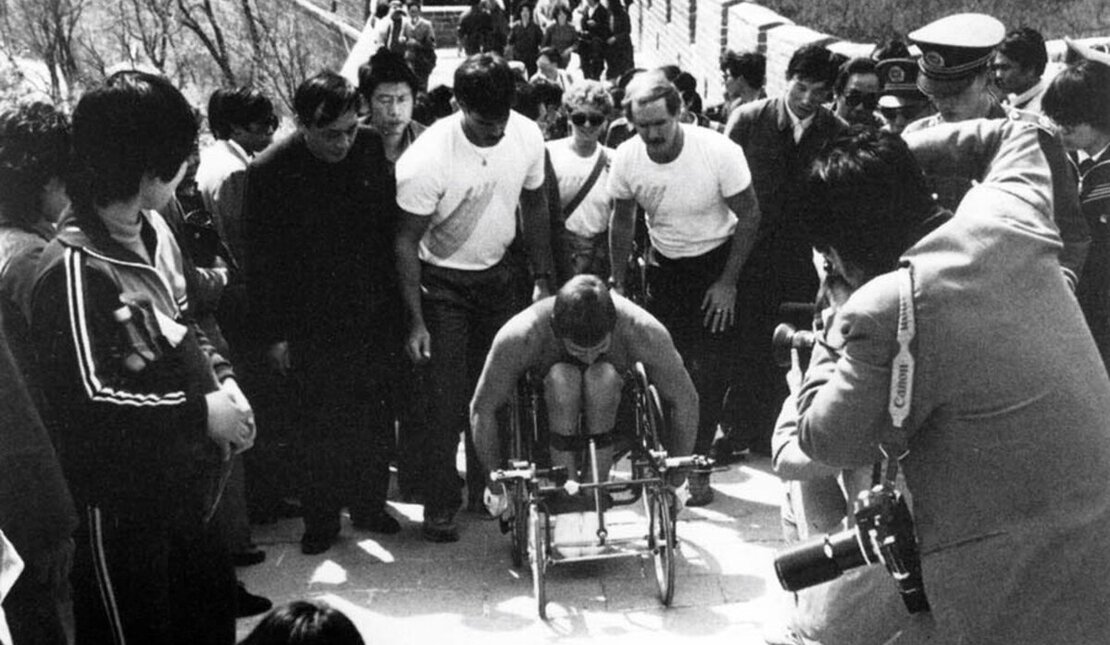 Black and white photo of Rick Hansen surrounded by a crown of people as he pushes up the Great Wall of China.
