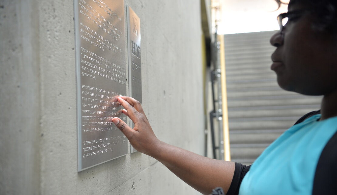 Woman wearing a teal shirt reading a metal braille sign that is mounted on a wall.