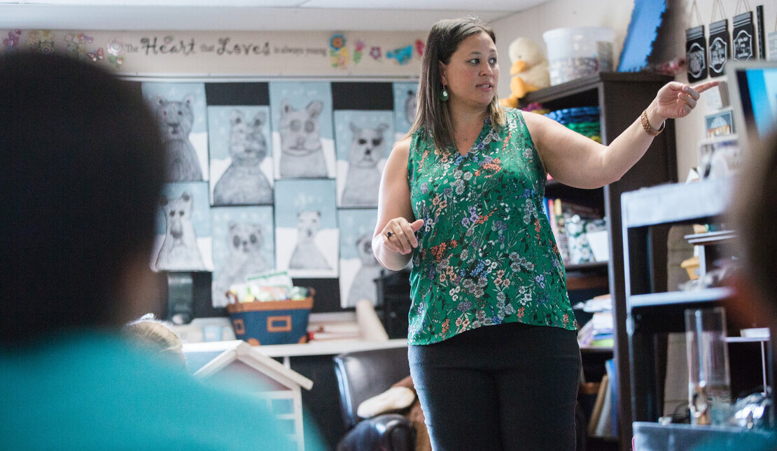 Teacher with long brown hair wearing a green shirt speaking in the front of a classroom