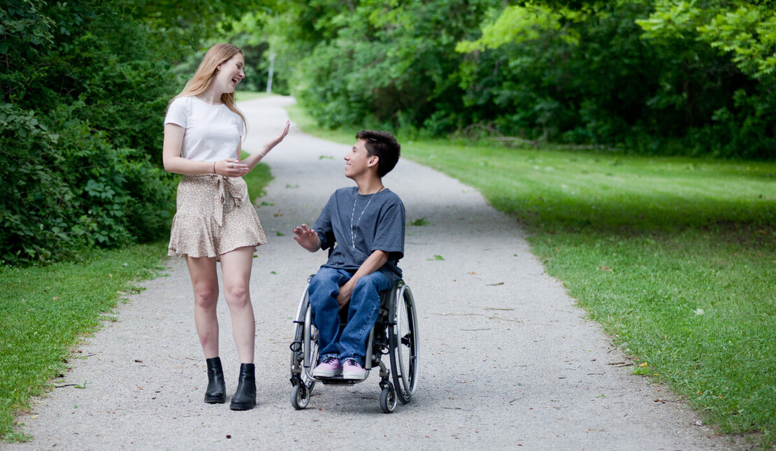 Two people talking and laughing while on a gravel path. One is using a wheelchair, has short brown hair and is wearing a dark t-shirt. The other person has long hair, is wearing a skirt and is walking.