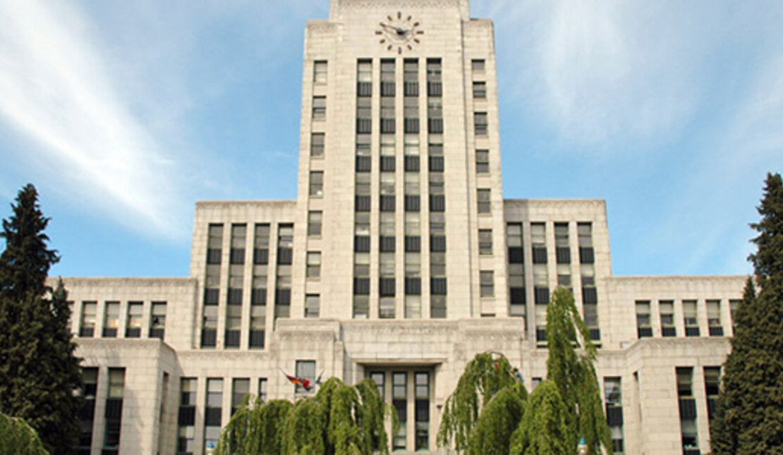 Vancouver City Hall, a large beige building with a clock at the top. It is surrounded by trees.