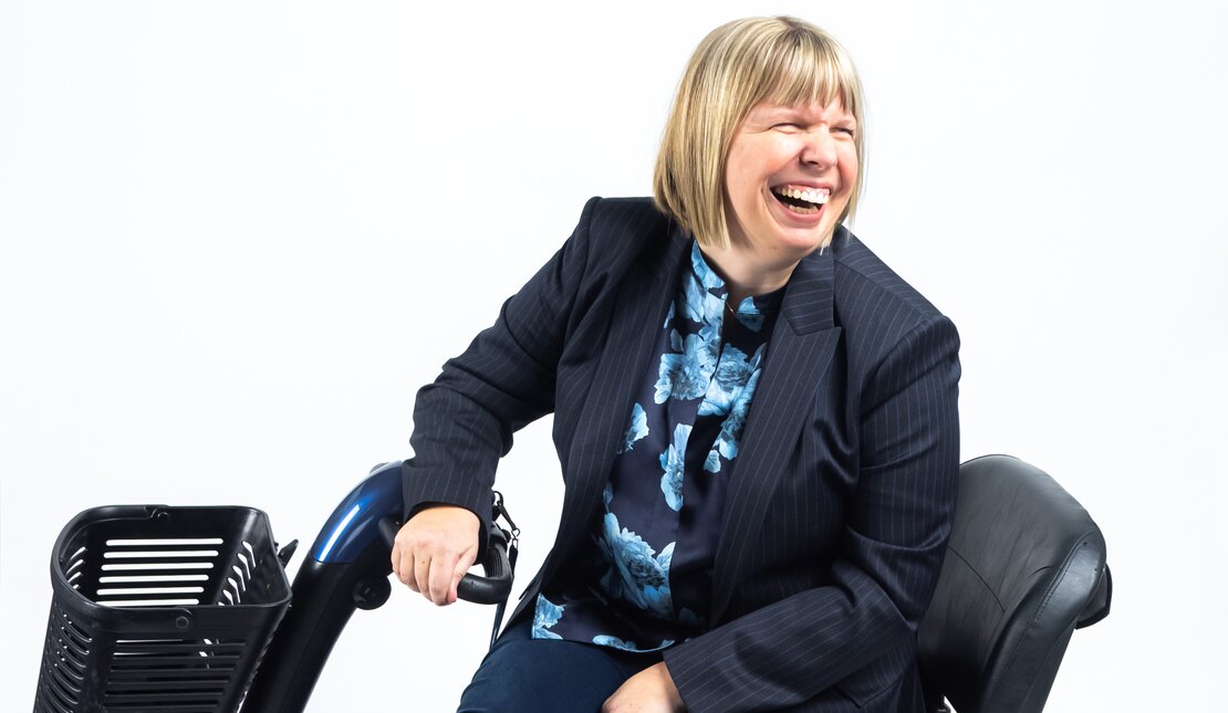 Darby Lee Young who has short blonde hair cut in a bob. Darby is wearing a striped blazer, a floral shirt and shiny blue shoes. She is laughing and is using a motorized mobility device.