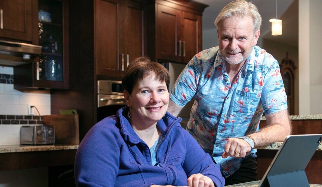 Couple at their kitchen counter which is at an accessible height. There is an iPad on the counter. The man has grey hair and is standing next to his wife who has short brown hair and is using a wheelchair.