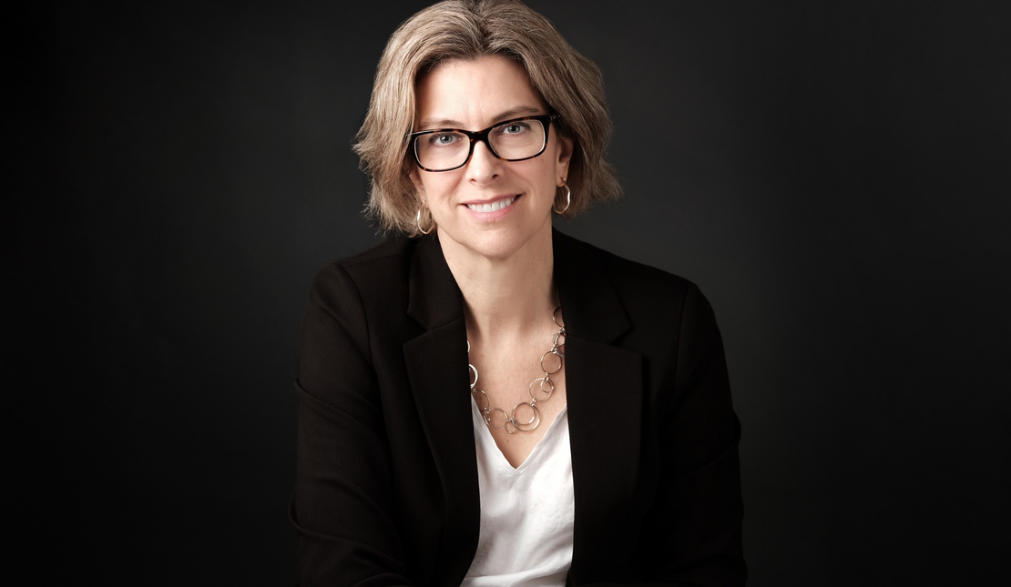 Headshot of Stephanie Cadieux who has short brown hair and glasses. She is wearing a black blazer and white shirt and is in front of a black background.