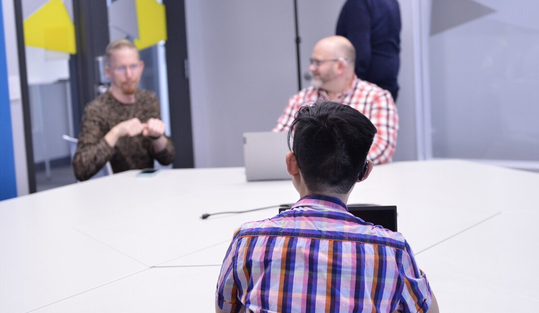 Back of the head of someone with short dark hair wearing a plaid shirt, has a laptop open and sitting at a table with two other people. One person is using American Sign Language