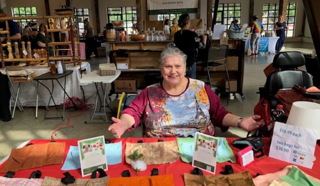 Julie Melanie, who has grey hair that is in a pony tail, sitting at a table with an orange table cloth that has her bags on display.