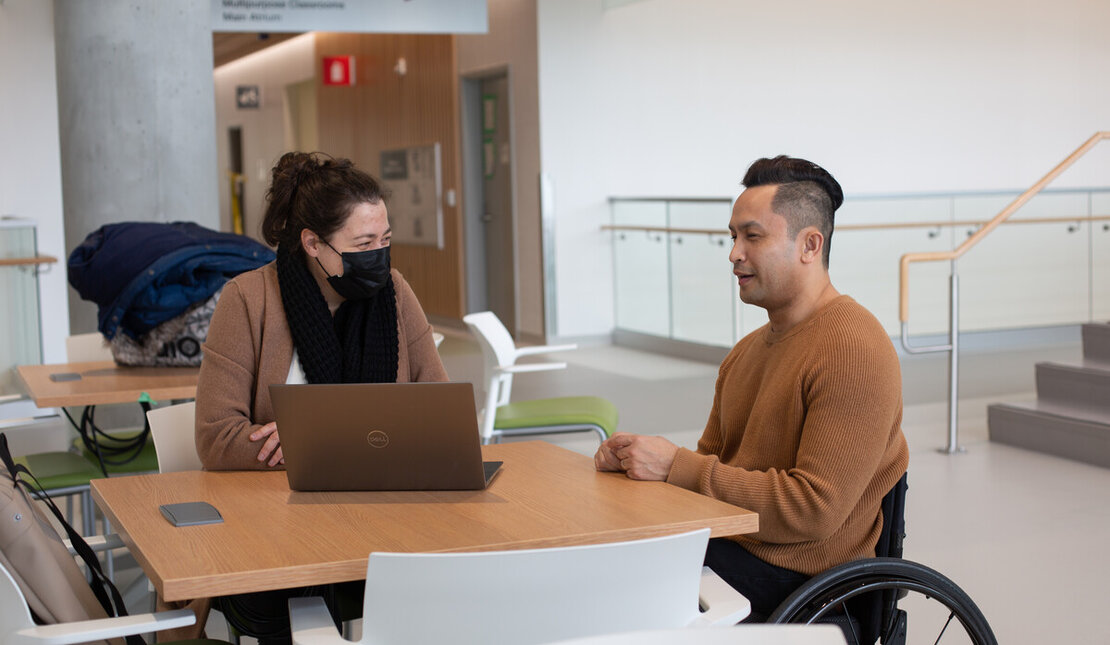 Two people working at a desk using a computer. One person is wearing a face mask and the other person is using a wheelchair.