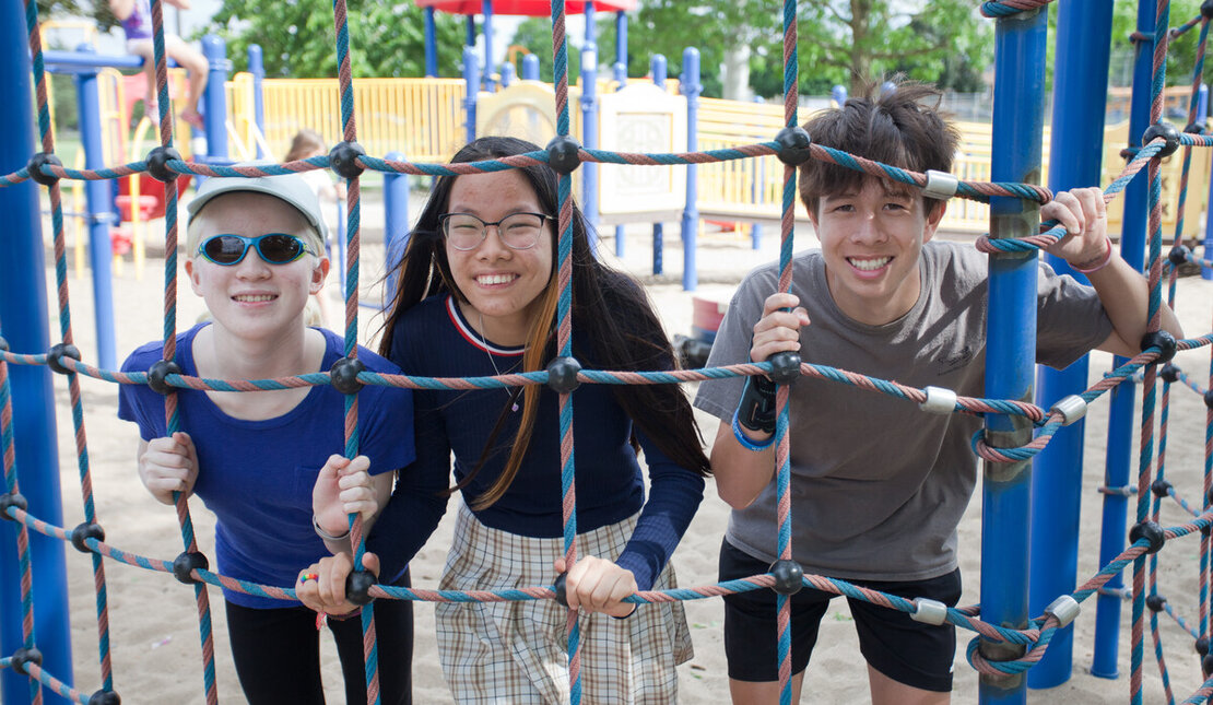 Three young people on a playground smiling and looking through a climbing rope. One youth has low vision and is wearing sunglasses.