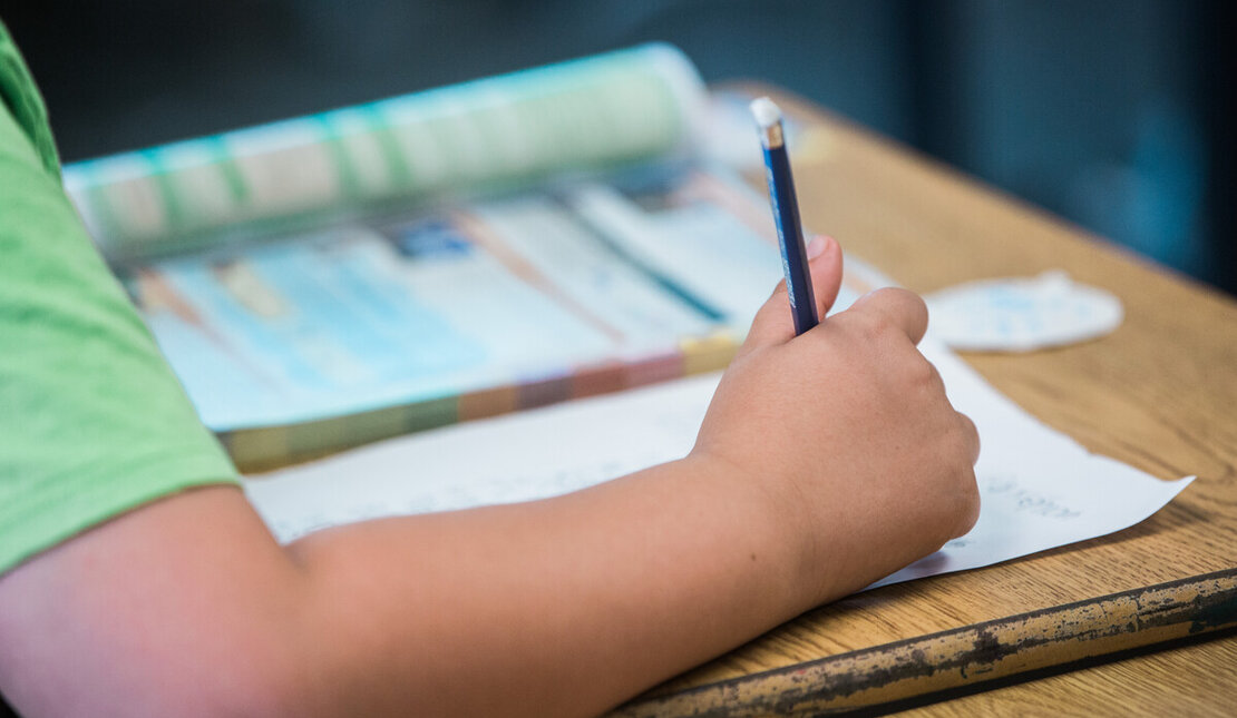 A child's arm writing on a piece of paper with a pencil. There is an open book on the desk.