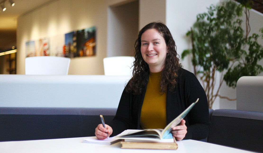 Amanda Motyer sitting at a table with an open book and pen. She has curly brown hair and is wearing a brown shirt and black blazer.