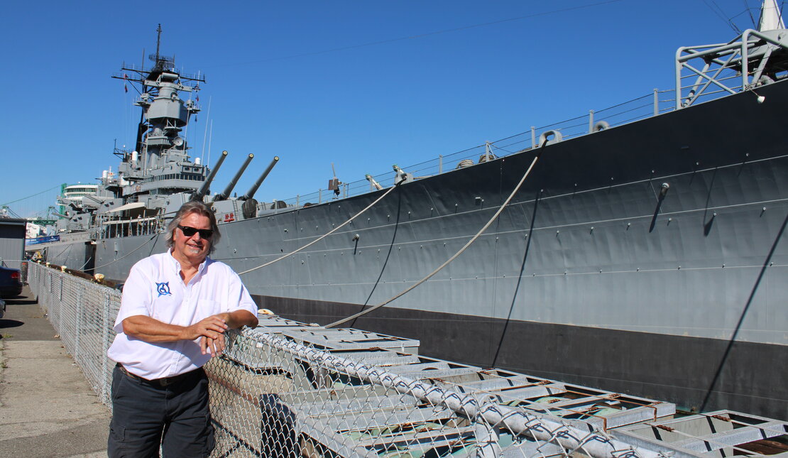 Colin McCarthy leaning on a chain link fence next to a large military ship. Colin has chin-length grey hair and is wearing a white shirt and black shorts.