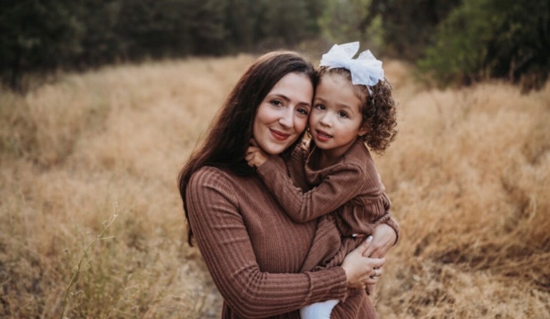 Katie holding her daughter Holland. They are wearing matching brown sweaters and are in front of a field.