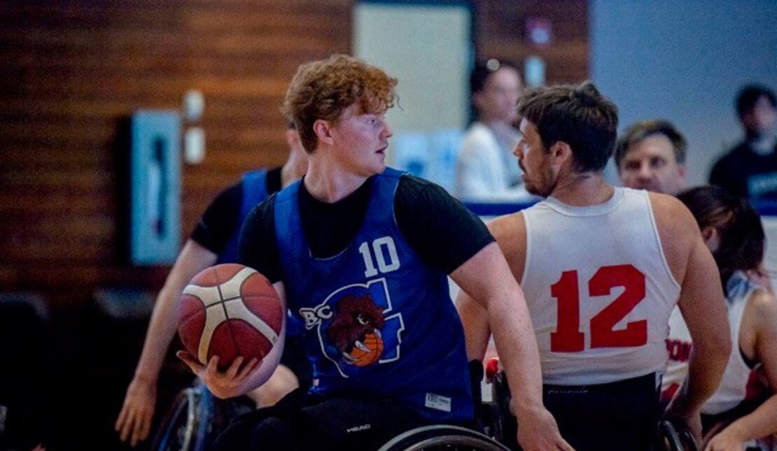 Cameron playing wheelchair basketball. He is holding the ball and looking back at an opponent. 