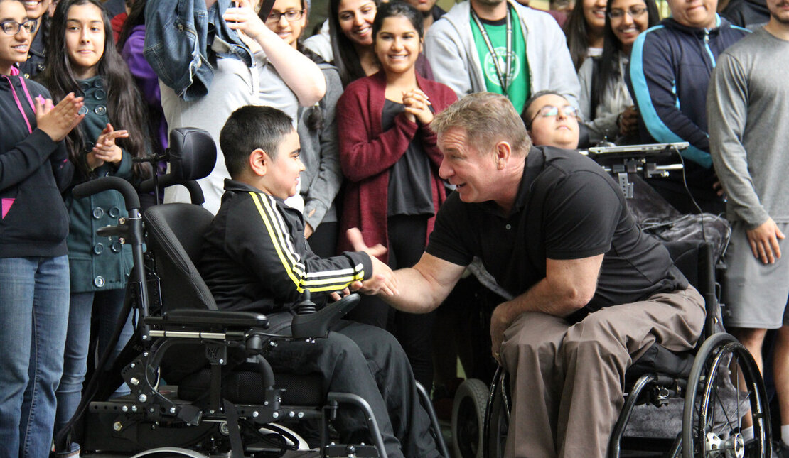 Rick Hansen shaking the hand of a young person who is using a wheelchair. A large crowd of students cheers behind them