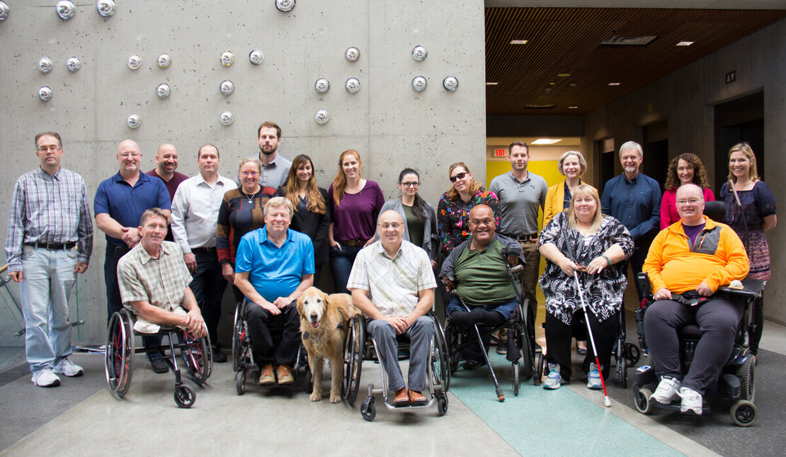 A group of people with different abilities smiling in front of a grey wall.