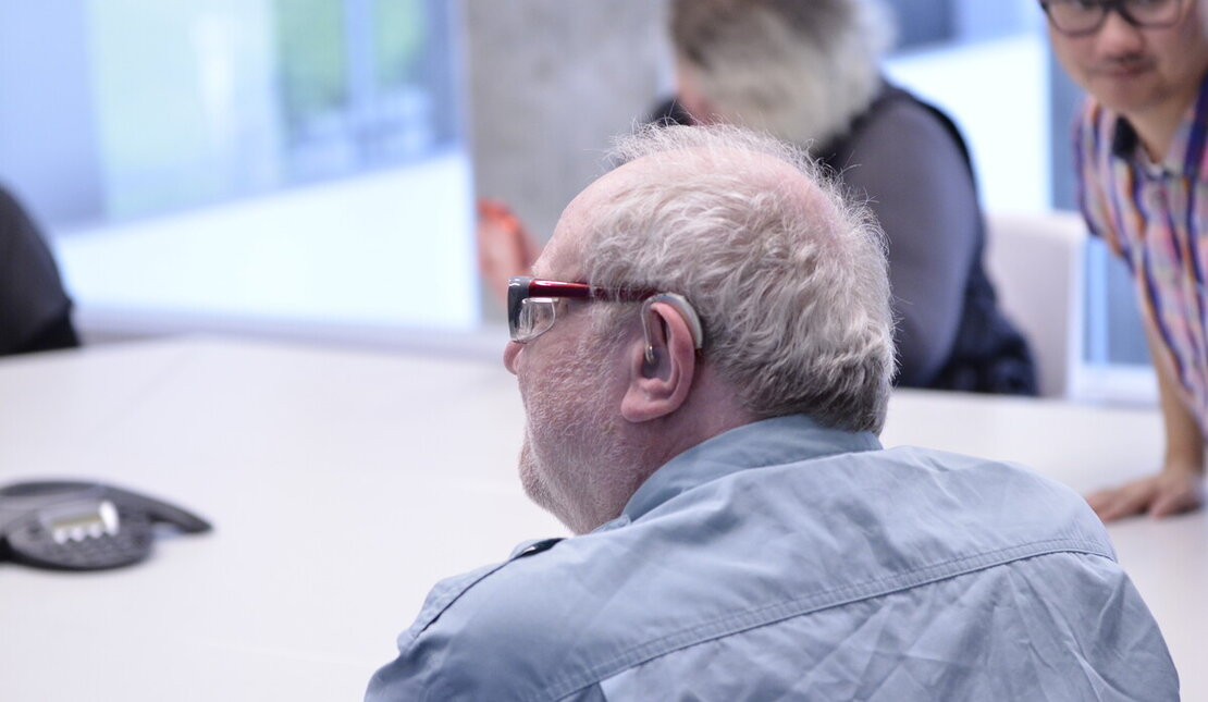 AN elderly man who is wearing glasses and has a hearing aid. He is sitting at a table with others.
