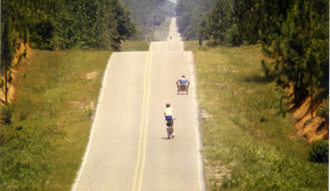 Rick Hansen during the Man In Motion World Tour. He is in the distance wheeling up a very large hill. A person on a bicycle rides behind him.