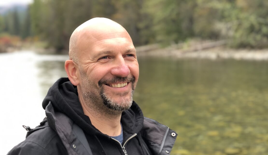 A man with a salt and pepper coloured beard by a river. He is wearing a black sweater and rain jacket and smiling.