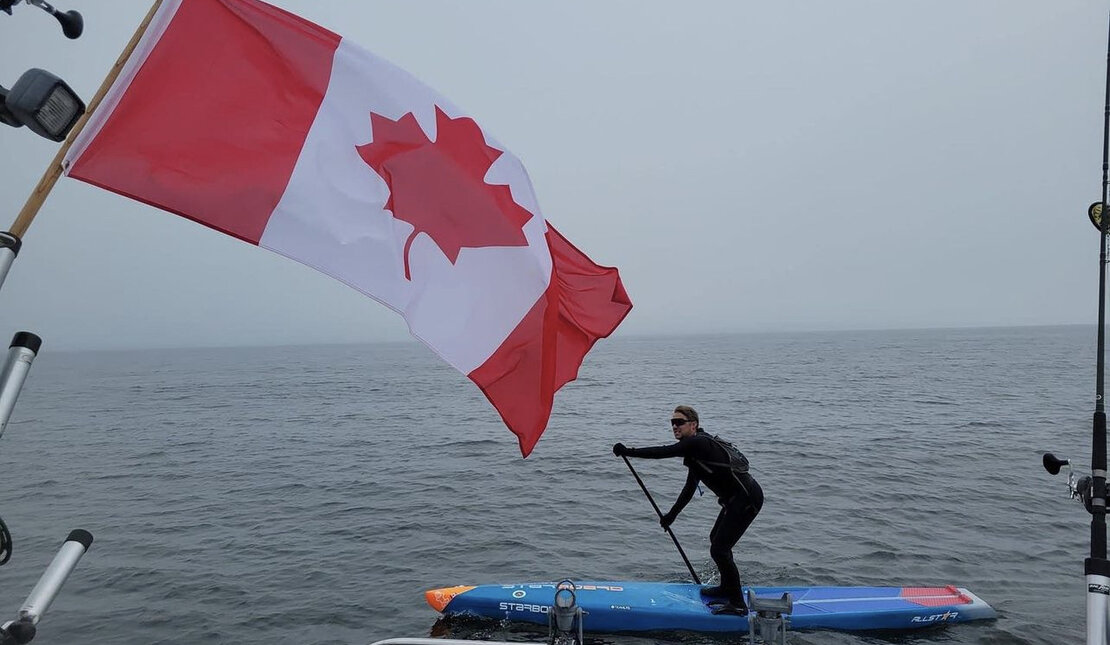 Mike Shoreman on a paddleboard in the lake. There is a large Canada flag in front of him
