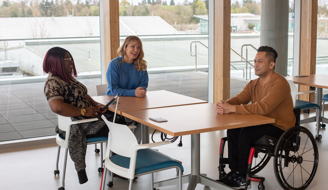 A woman with a white cane, a woman with a hearing aid and a man in a wheelchair sit around a table