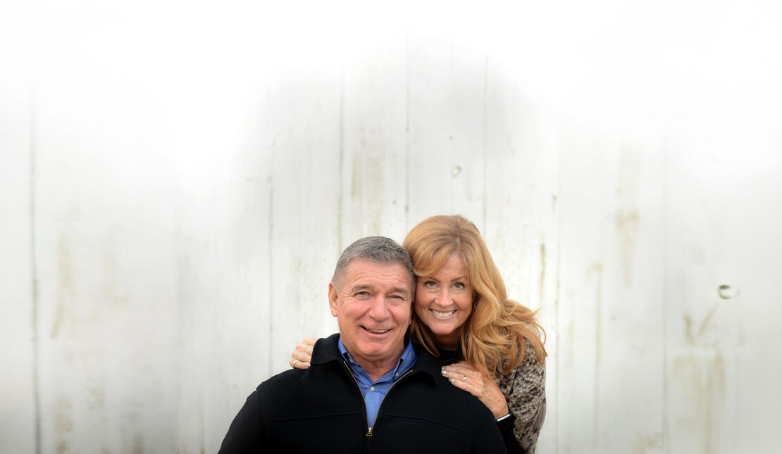 Rick and Amanda Hansen, smiling against a white backdrop, with Amanda hugging the Rick lovingly from behind.
