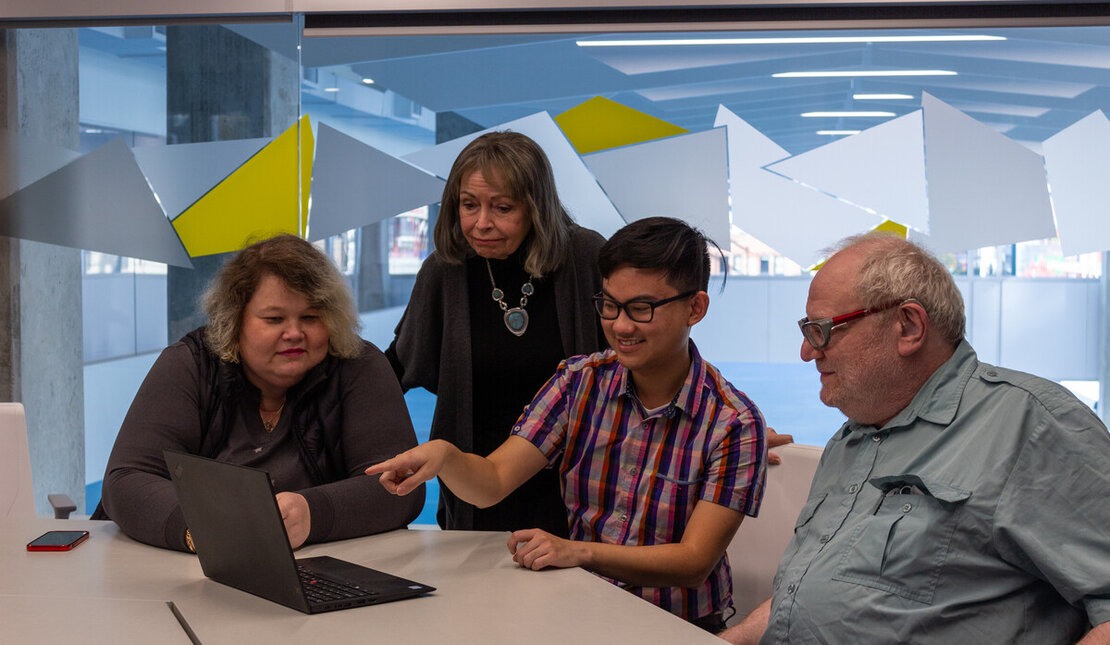 four people in professional attire look at a laptop in a boardroom