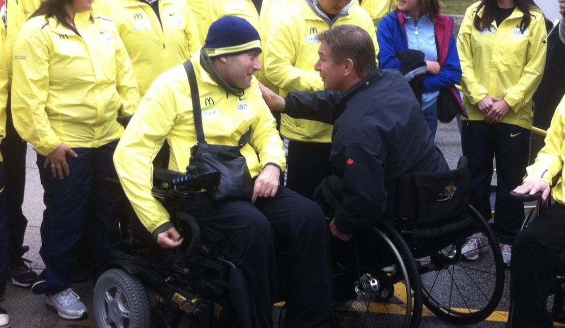 Two men in wheelchairs talking and smiling at each other, surrounded by people