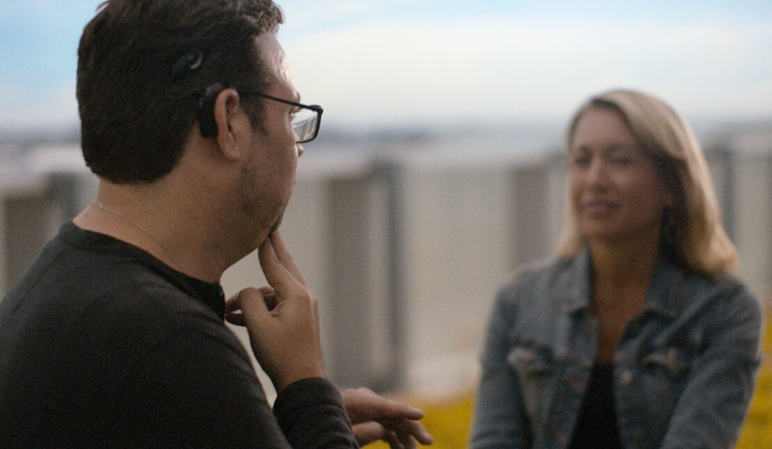 A man with is shown in profile, gesturing near his face while speaking to another person in sign language. They are outdoors, with a fence and greenery visible in the background.
