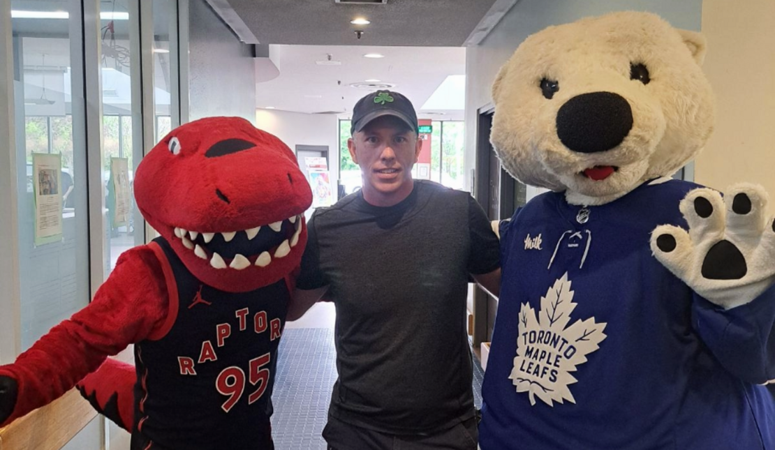 An individual standing with their arms around two sports mascots. On the left, there is a red dinosaur mascot wearing a black Toronto Raptors basketball jersey. On the right, there is a white bear mascot wearing a blue Toronto Maple Leafs hockey jersey.
