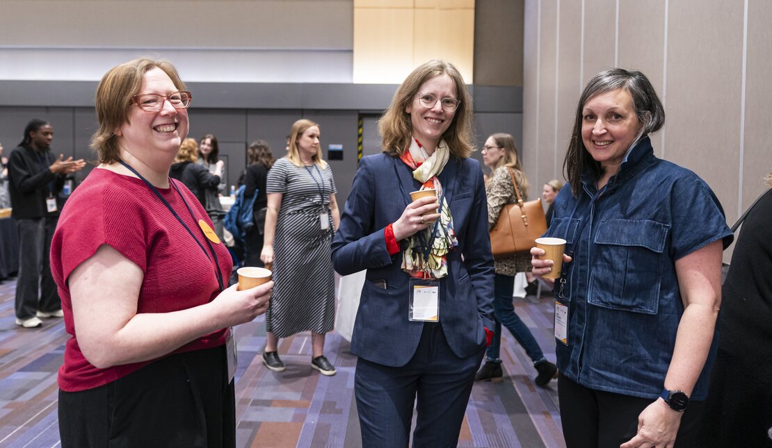 Three middle age woman standing  and holding a paper coffee cup. They are engaged in conversation