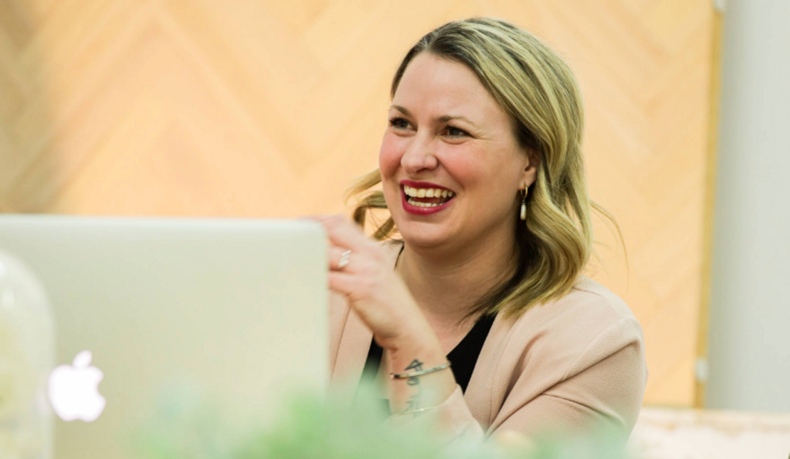 A woman seated at a desk with an open laptop in front. She is smiling and has one hand is raised near the face.