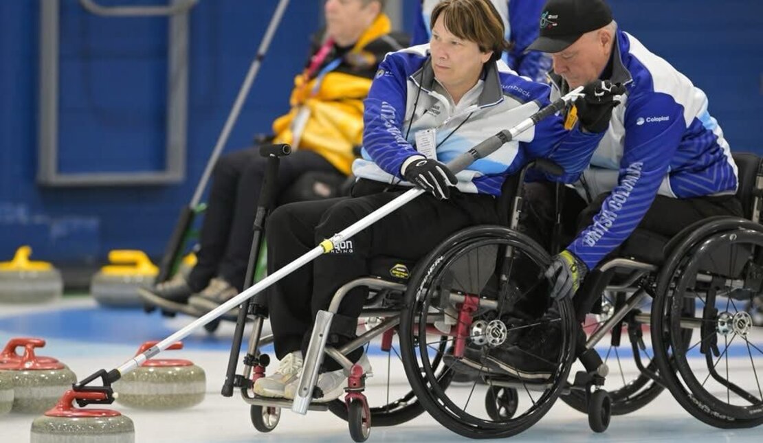 Two wheelchair curlers on the ice, both wearing team jackets and holding delivery sticks to push curling stones. 