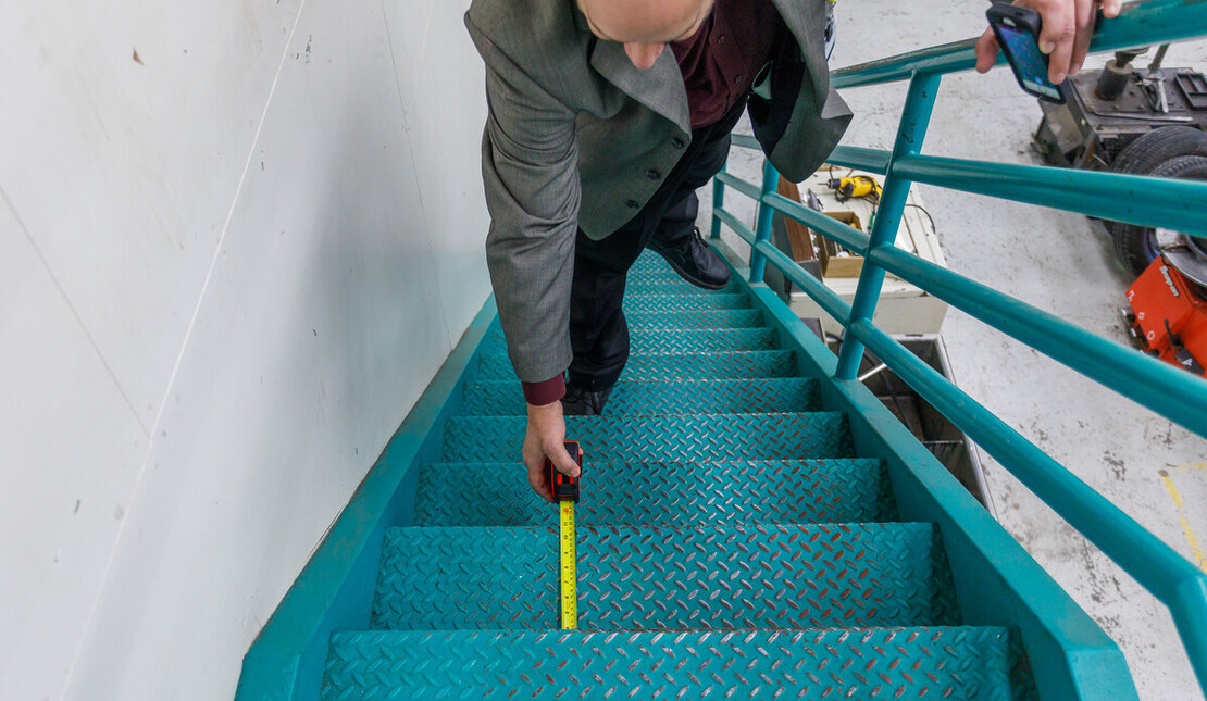 Person measuring the depth of teal metal stairs using a yellow tape measure. The stairway has textured anti-slip surfaces and teal railings
