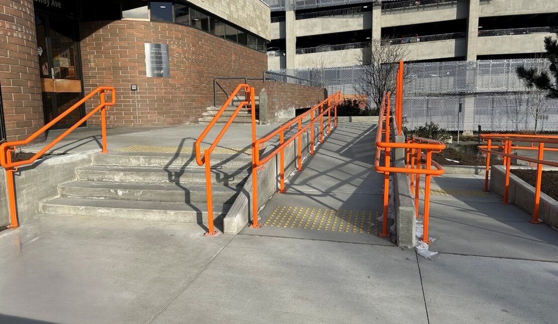 Concrete entrance with stairs and a ramp featuring bright orange handrails and tactile warning strips leading to a museum door.