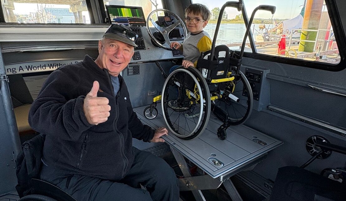 Inside a boat cabin, Rick Hansen gives a thumbs-up while a kid sits in a wheelchair near the steering wheel.
