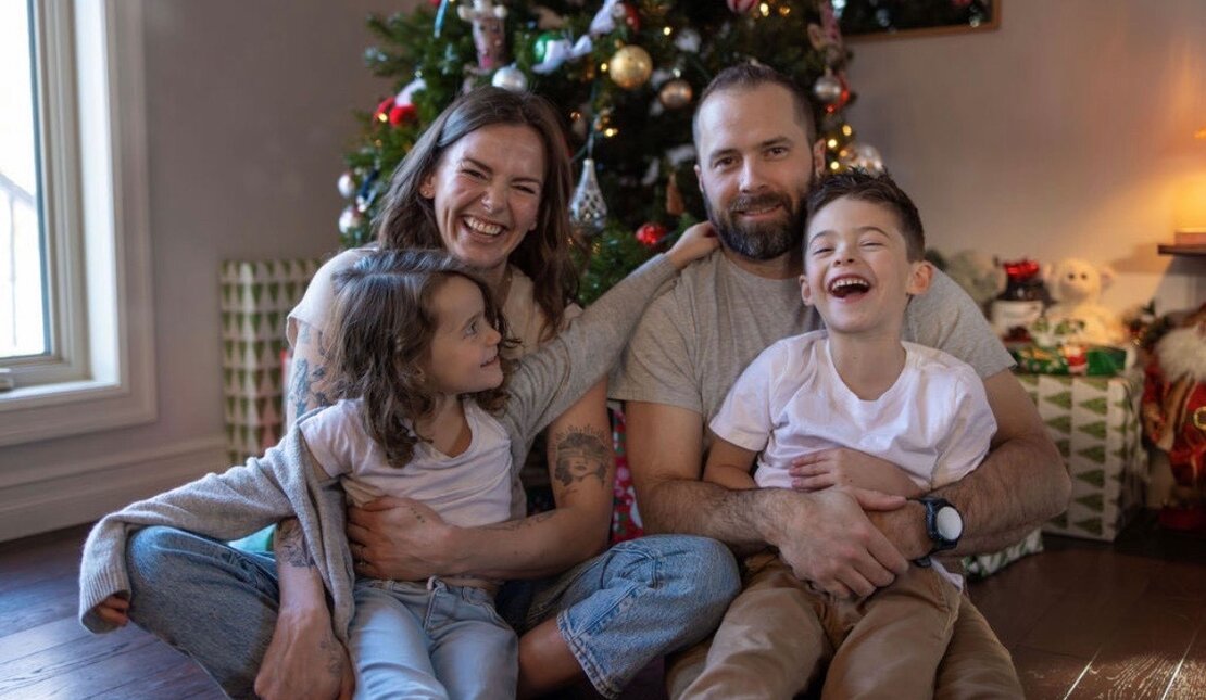 A family of four sits together on the floor, two children laughing in front of a decorated Christmas tree.
