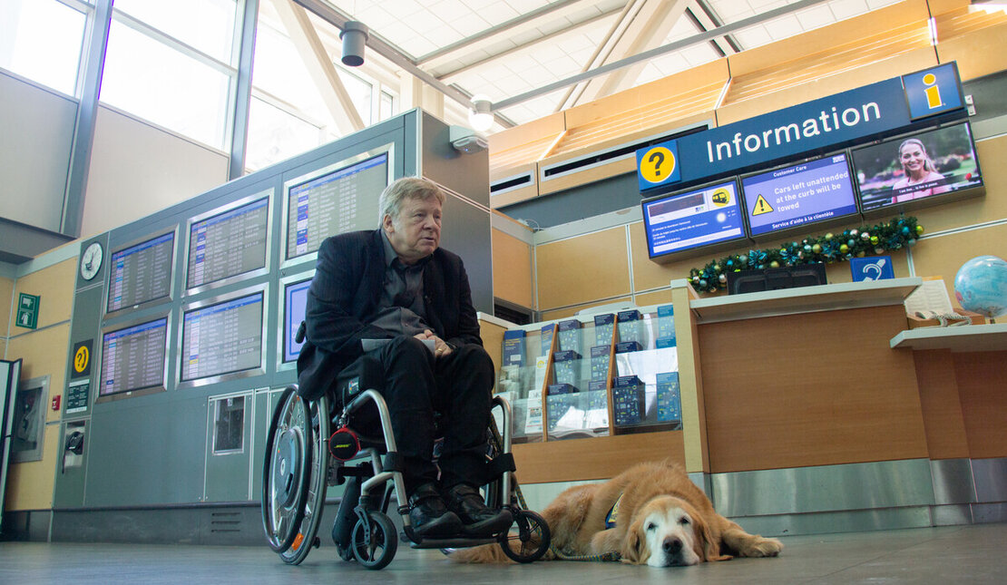 Person using a wheelchair waits at the aiport while a guide dog lies on the floor nearby, with information screens visible in the background.