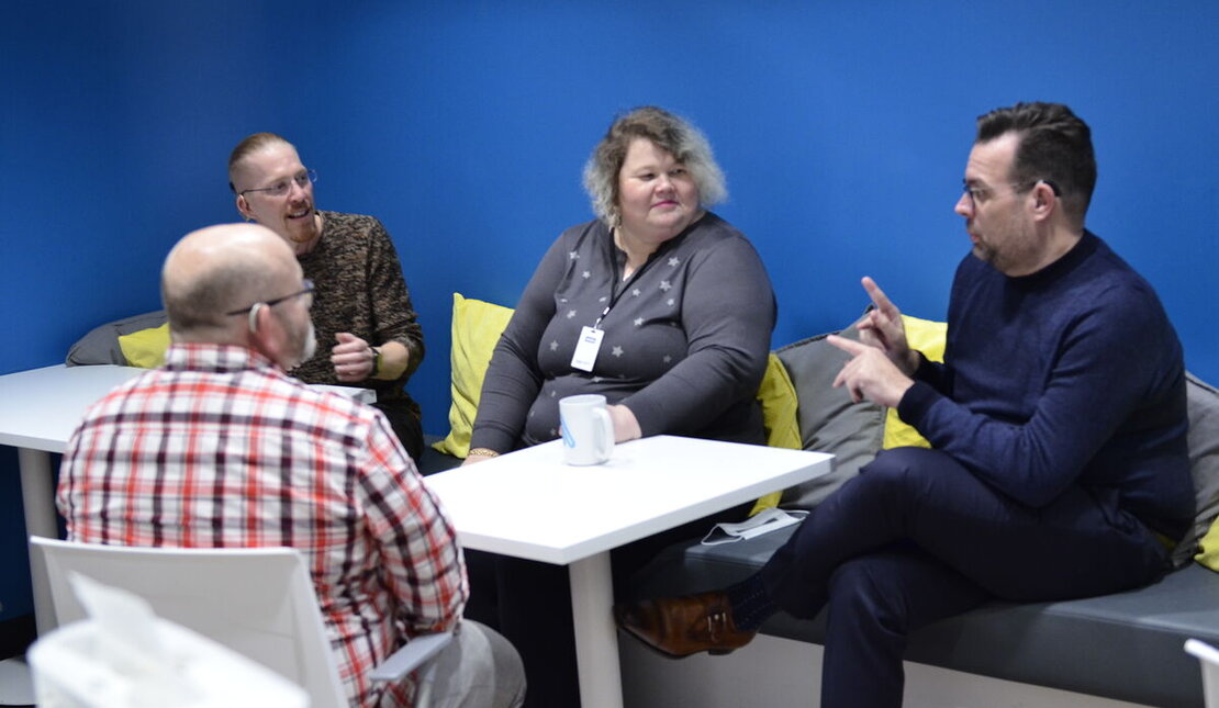 Four people of different ages seated around a small table in a casual conversation. One of them is wearing a hearing aid