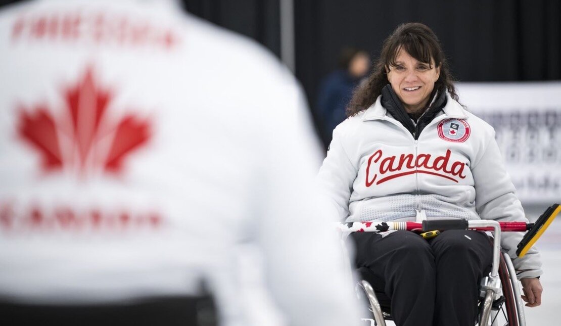 An athlete woman using a wheelchair in a curling ice rink with white background wearing a short‑sleeve athletic shirt with sports logos.
